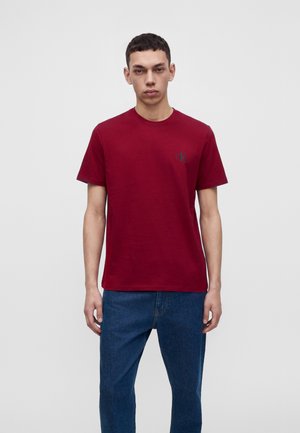 Jeune homme portant un t-shirt uni rouge foncé à manches courtes et un jean bleu, debout sur un fond uni gris clair.
