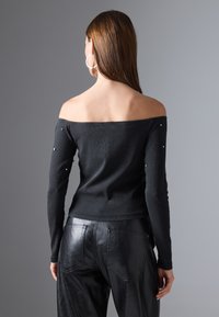 Hosszú barna hajú nő fekete, vállnélküli blúzt visel, gyöngyökkel díszített ujjal és fekete bőr nadrággal, háttal áll a kamerának.