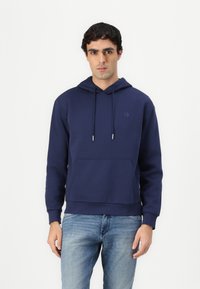 Navy blue hoodie in soft fabric with long sleeves, a front pocket, drawstring hood, and subtle logo detail on the chest. Paired with light jeans.