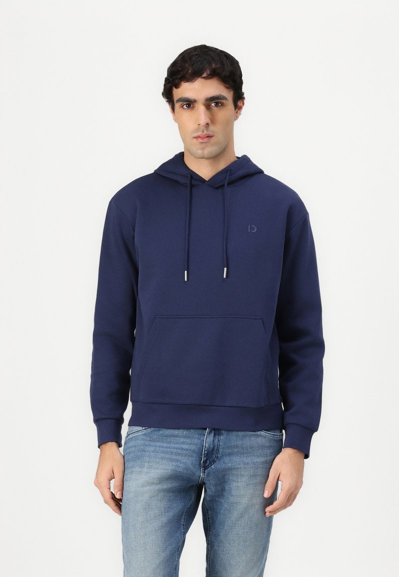 Navy blue hoodie in soft fabric with long sleeves, a front pocket, drawstring hood, and subtle logo detail on the chest. Paired with light jeans.
