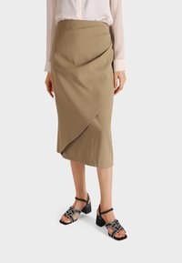 Gonna midi beige con un design drappeggiato, realizzata in tessuto morbido. Abbinata a sandali neri con tacco a blocco dotati di cinturini decorativi.