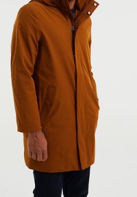 Ruskea vedenkestävä takki, jossa on huppu, edessä painonapit ja sivutaskut. Sileä pinta ja suora, keskipitkä leikkaus.