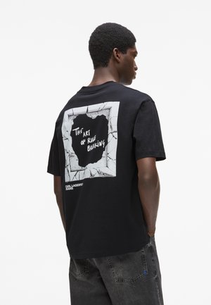 Homme portant un t-shirt noir avec un graphisme de mur fissuré et le texte "L'art de transgresser les règles" au dos, assorti à un jean gris foncé.