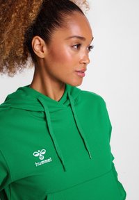 Grüne Kapuzenjacke aus Baumwollmischung, mit einer Fronttasche, verstellbarem Kordelzug an der Kapuze und weißem "hummel"-Logo auf der Brust.