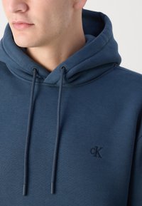 Dunkelblaues Hoodie mit Kordelzug, aus weichem Stoff gefertigt. Verfügt über ein gesticktes Logo auf der Brust und eine strukturierte Kapuze.