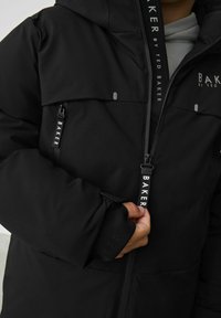 Schwarze Außenjacke mit Reißverschluss, zwei Vorderaschen und Textbranding. Der Stoff hat eine glatte Textur mit matter Oberfläche.