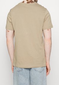 Béžové bavlněné tričko s kulatým výstřihem, krátkými rukávy a volným střihem, pohled zezadu s jemnými skvrnami na látce.