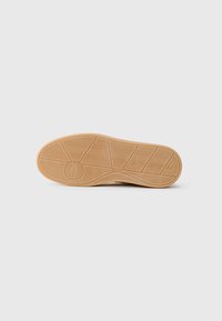 Semelle d'une chaussure en caoutchouc marron clair avec un motif de semelle texturé pour une meilleure adhérence, et un logo circulaire au centre.