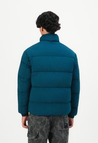 Hombre con cabello rizado y oscuro que lleva una chaqueta acolchada color teal y pantalones cargo grises con camuflaje, de pie frente a un fondo blanco liso.