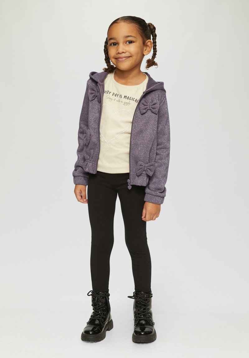 Lila Kapuzenpullover mit Schleifen-Details, cremefarbenes Shirt mit Aufdruck, schwarze Leggings und schwarze Schnürstiefel. Glatte Texturen, lässiges Outfit.
