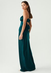 Teal satiini iltapuku, jossa on tiukka bodice, rypytetty selkä ja sivuaukot. Hame virtaa lattiaan sileällä tekstuurilla.