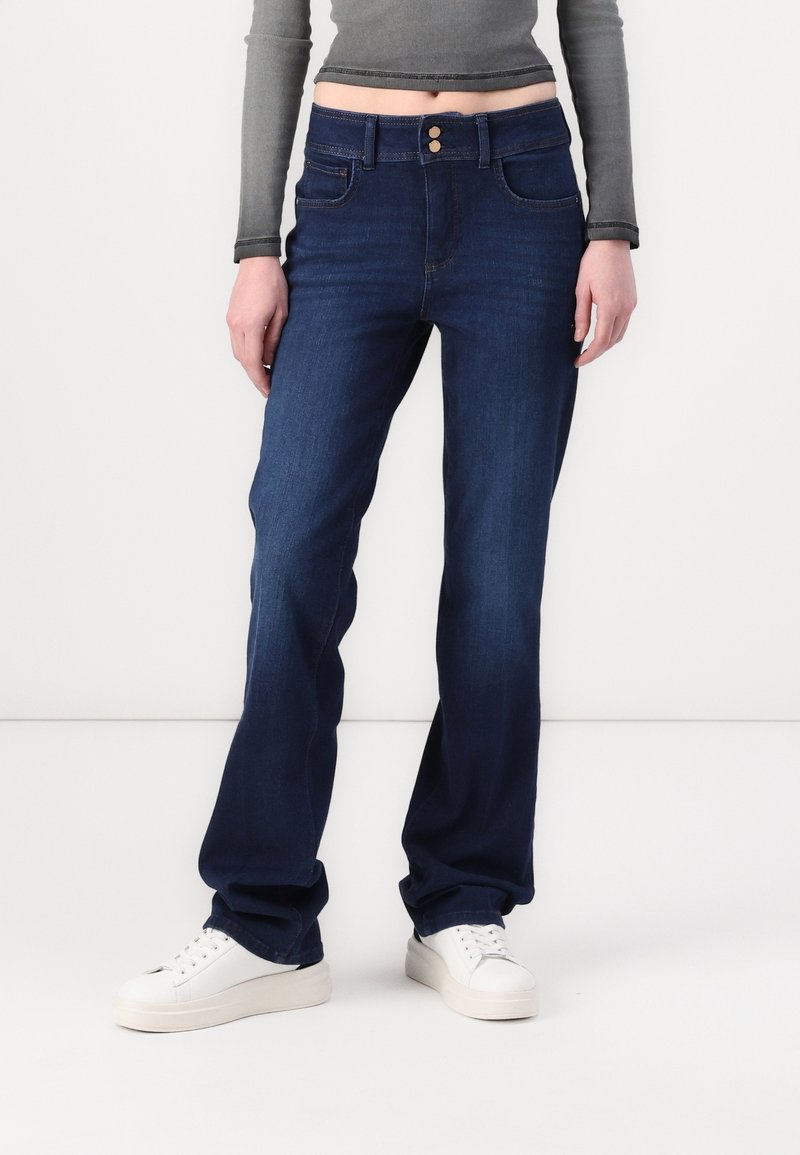 Personne portant un jean droit bleu foncé, des baskets blanches et un top court gris à manches longues, debout devant un fond blanc.
