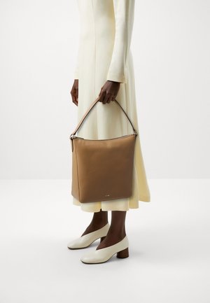 Sac tote en cuir brun avec un design structuré, fermeture éclair sur le dessus et une courte poignée. Modèle portant une longue robe couleur crème et des chaussures à talons blocs blancs.