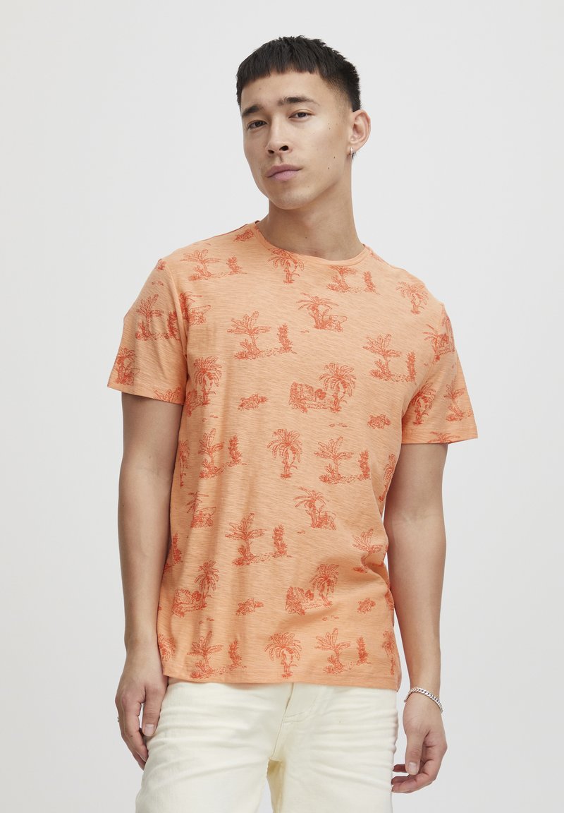 Kortermet oransje t-skjorte laget av teksturert stoff, med et gjentatt trykk av palmer og natur i mørkere oransje nyanser.