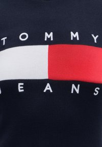 Ναυτικό ύφασμα με το κείμενο «TOMMY JEANS» πλεγμένο πάνω και κάτω από ένα οριζόντιο ορθογώνιο μπλοκ βαμμένο λευκό αριστερά και κόκκινο δεξιά.