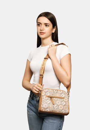 Giovane donna con t-shirt bianca e jeans che tiene una borsa a tracolla beige a fantasia con tracolla regolabile sulla spalla contro uno sfondo semplice.