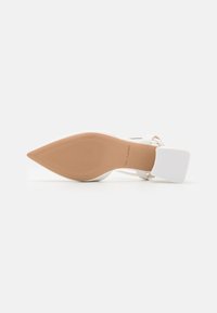 White high-heeled shoe with a pointed toe, thin straps, and a square heel. The sole is beige with a textured pattern, featuring brand lettering.