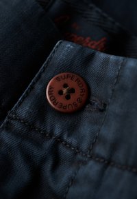 Donkerblauwe denim stof heeft een bruine knoop met het ingegraveerde woord "SUPERDRY" en vier gaten, gelegen nabij een naad op het kledingstuk.
