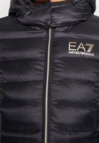Svart vadderad jacka med dragkedja framtill, med quiltade sektioner, hög krage och en EA7 Emporio Armani-logotyp i guld på vänster bröst.
