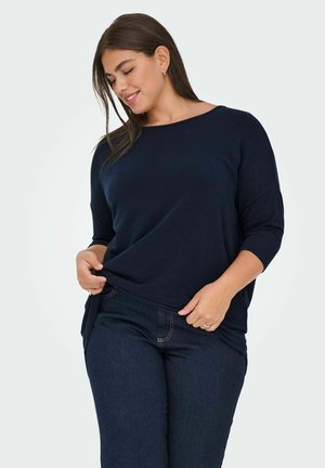Donna con lunghi capelli scuri, che indossa una maglia a maniche lunghe blu navy e jeans scuri, sorridente mentre tiene l'orlo della maglia.