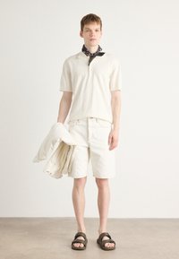 Polo shirt beige clair à manches courtes, shorts assortis et une légère veste. Écharpe noire à motifs nouée autour du cou. Sandales marron avec boucles.