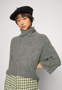 Sivi pulover s rolkragnom, labavog kroja i kratkim rukavima, izrađen od teksturirane pletene tkanine, uparen s houndstooth uzorkom zelenih hlača.