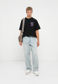 Black t-shirt with a purple mountain graphic pocket, light blue baggy jeans, and white sneakers with grey accents, carrying a grey shoulder bag.