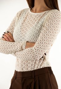 Top in pizzo a maniche lunghe color avorio con motivi geometrici e scollatura rotonda, abbinato a pantaloni marroni a vita alta.