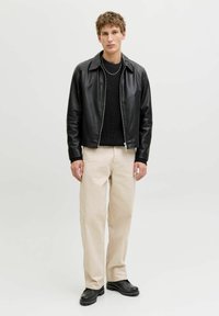 Młody mężczyzna stoi, ubrany w czarną skórzaną kurtkę, czarny sweter, beżowe spodnie, czarne buty i srebrny naszyjnik, na jednolitym tle.