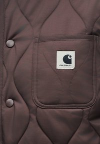 Καφέ γιλέκο με καπιτονέ σχέδιο που διαθέτει τσέπη στο στήθος με ασπρόμαυρο λογότυπο. Περιλαμβάνει στρογγυλά καφέ κουμπιά και υφασμάτινη υφή.