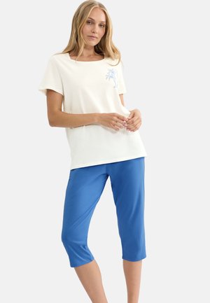 Kobieta w białej bluzce z krótkim rękawem i niebieskim kwiatowym wzorem oraz niebieskich skróconych spodniach, stojąca ze splecionymi rękami na wysokości talii.