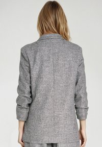 Szary żakiet wykonany z fakturowanej tkaniny, charakteryzujący się dopasowanym krojem, klapami i rękawami do uda. Widok z tyłu ukazuje staranne wykończenie.
