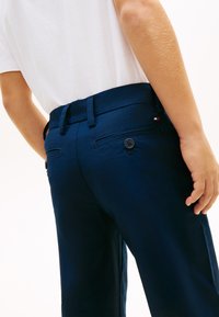 Άτομο που φοράει σκούρο μπλε παντελόνι με τσέπες στο πίσω μέρος με κουμπιά και λευκό πουκάμισο μέσα από το παντελόνι, με το χέρι χαλαρό δίπλα στο σώμα.