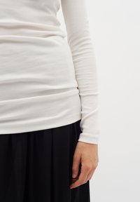 Vit långärmad ribbad topp med figurnära design, kombinerad med en svart plisserad kjol som visar upp textilstruktur och smidiga linjer.