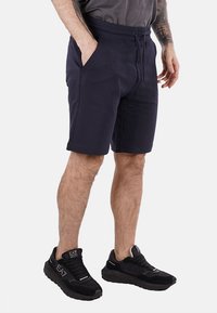 Bermuda blu navy realizzati in tessuto morbido, con un elastico in vita e cordoncino, e tasche laterali. Abbinati a scarpe da ginnastica nere.