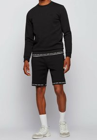 Conjunto de sudadera negra y pantalones cortos, con una banda de logo en el dobladillo. La sudadera tiene un escote redondo; los pantalones cortos son de longitud a la rodilla y ajustados.
