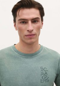 Lys grøn t-shirt lavet af bomuld, med rund halsudskæring og et grafisk print med et kranium og tekst på venstre bryst.