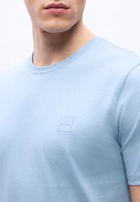 Άνδρας που φοράει γαλάζιο T-shirt με στρογγυλή λαιμόκοψη και μικρό ανάγλυφο λογότυπο BOSS στο στήθος, με φόντο λευκό.