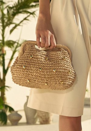 Mână care ține o geantă bej țesută, cu capse aurii, pe un fundal deschis cu plante și haine bej.