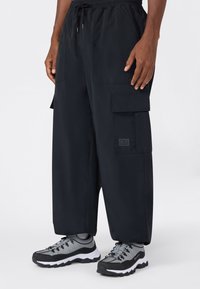 Persona vistiendo pantalones cargo anchos negros con bolsillos frontales y zapatillas deportivas grises y negras, de pie contra un fondo blanco.