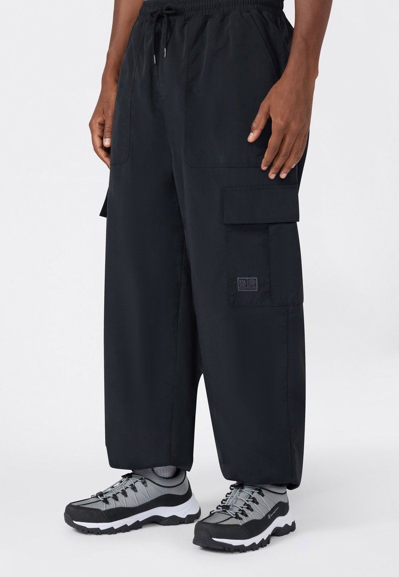 Persona vistiendo pantalones cargo anchos negros con bolsillos frontales y zapatillas deportivas grises y negras, de pie contra un fondo blanco.