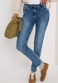 Blå denimjeans med en figurnära design, lätt blekning och sömmar, ihop med en tan atletisk sko och en olivgrön jacka.