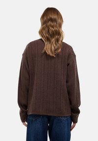 Pull en maille côtelée marron avec des manches longues, coupe ample et tissu texturé, associé à un jean en denim bleu taille haute.