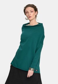 Turkusowa bluzka z długim rękawem, o asymetrycznym, drapowanym dekolcie i luźnym kroju. Posiada metkę z boku, zestawiona z czarną spódnicą. Miękka tekstura materiału.