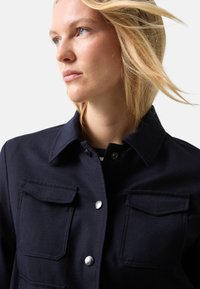 Blonde woman with blue eyes wearing a dark navy jacket with buttoned flap pockets, looking off to the side against a light background.