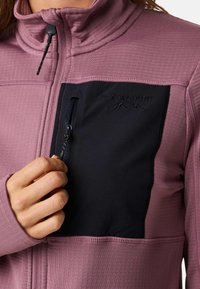Light pink full-zip jacket features a black chest pocket with a zipper, textured fabric, and contrasting color blocking. Logo embossed on pocket.