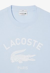 Camiseta unisex de algodón en azul claro con cuello redondo. Presenta un logo de cocodrilo blanco y el texto "LACOSTE PARIS" en impresión en negrita.