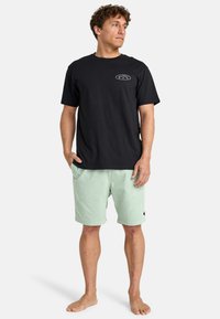 Sort T-shirt med korte ærmer og logo på brystet, parret med mintgrønne shorts, der har lommer og en tekstureret overflade. Ingen fodtøj.