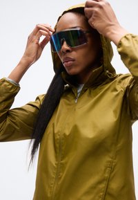 Chaqueta impermeable verde oliva con capucha; cuenta con una cremallera frontal y puños elásticos. La persona lleva gafas de sol oscuras y reflectantes.