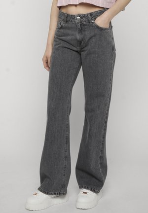 Persona che indossa jeans grigio scuro larghi e sneakers bianche con plateau, in piedi con una mano in tasca davanti a uno sfondo semplice.